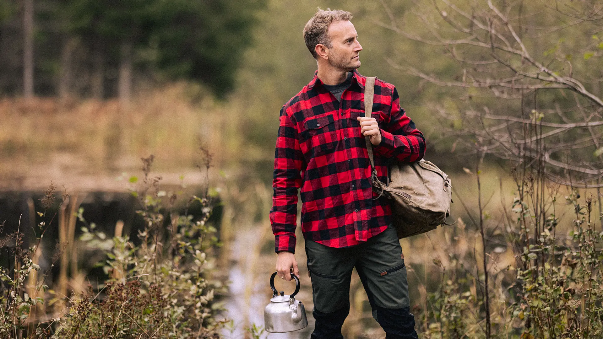 Mann ikledd rød flanellskjorte står med en gammel sekk og en kaffekjele og skuer utover skogen